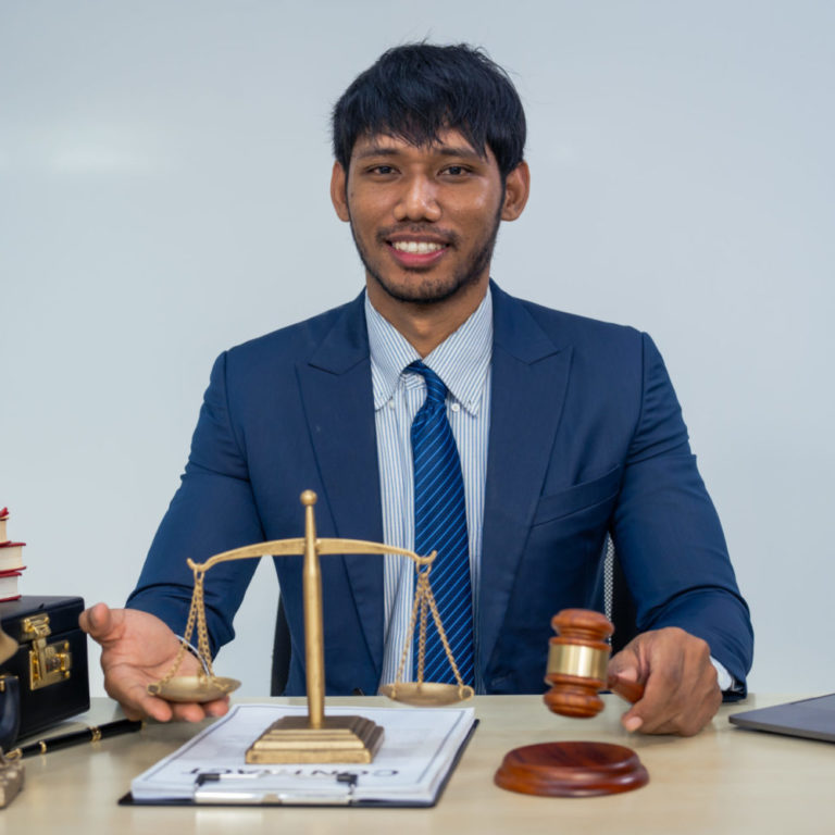 An Asian businessman in a formal suit works alone at a lawyer's desk, by golden scales, law books